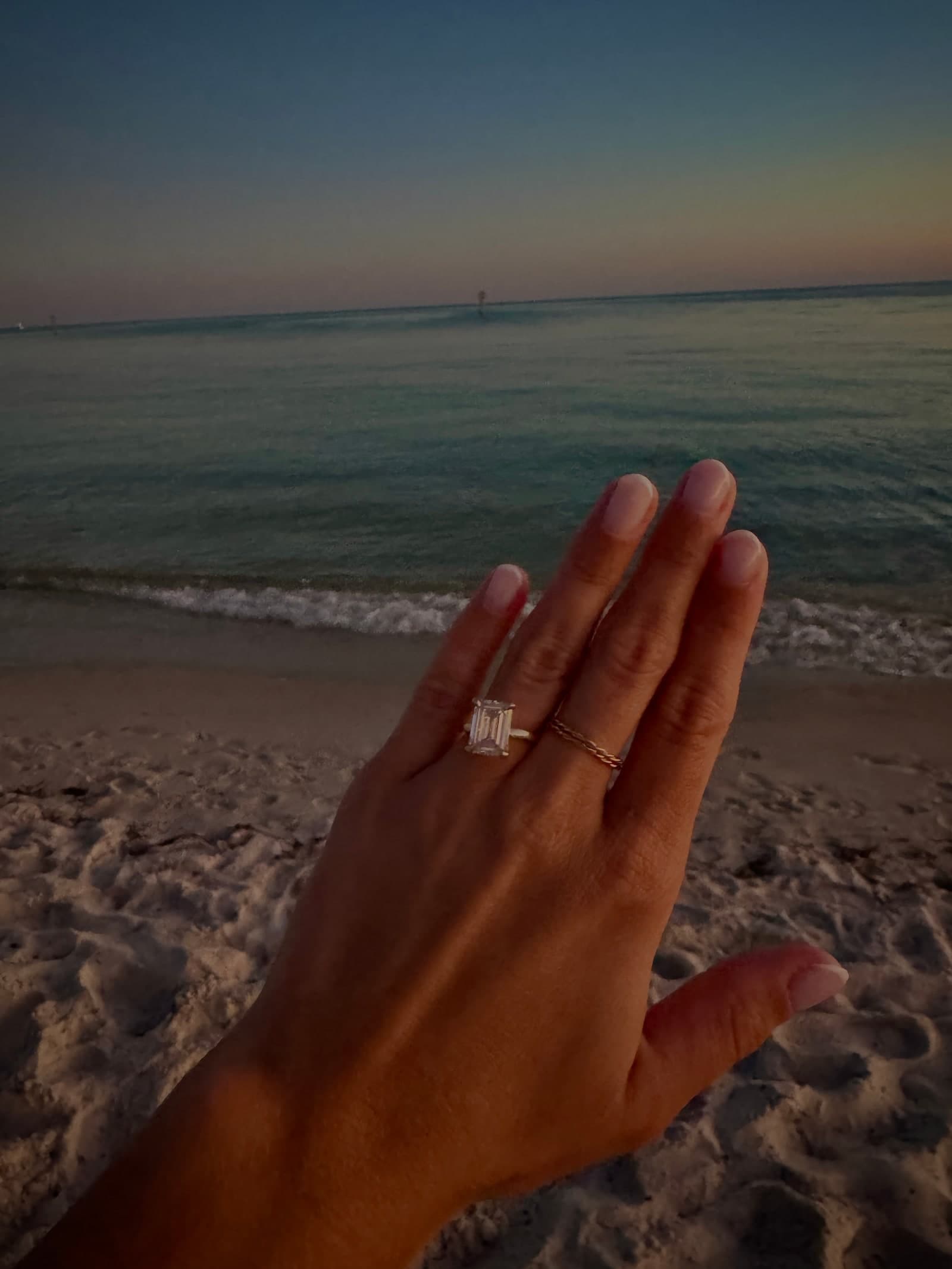 Hand wearing an emerald-cut engagement ring at sunset on the beach
